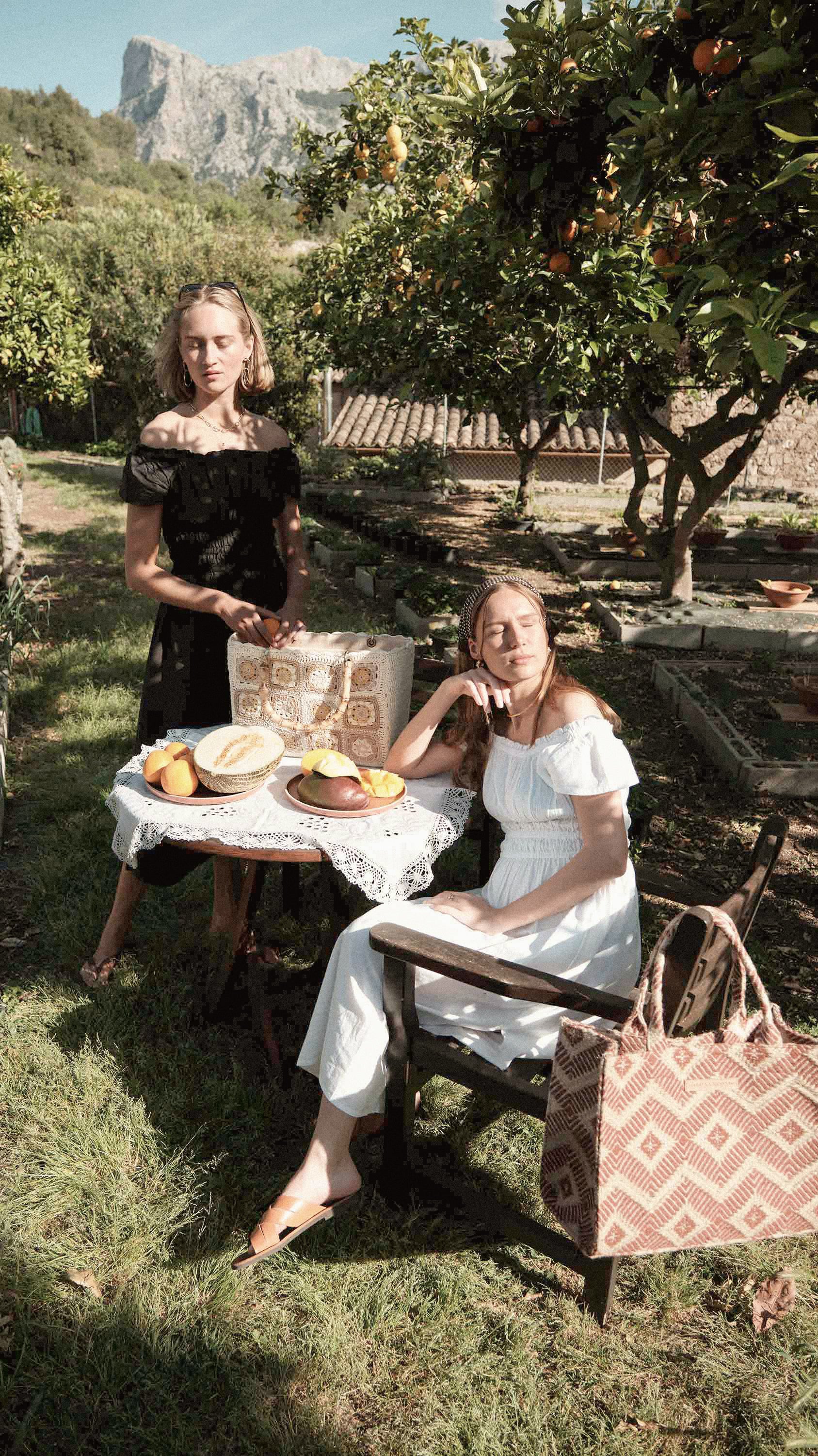 the two girls are wearing the positano off-the-shoulder dress in both black and white, paired with the la rambla bag in jute terracotta.